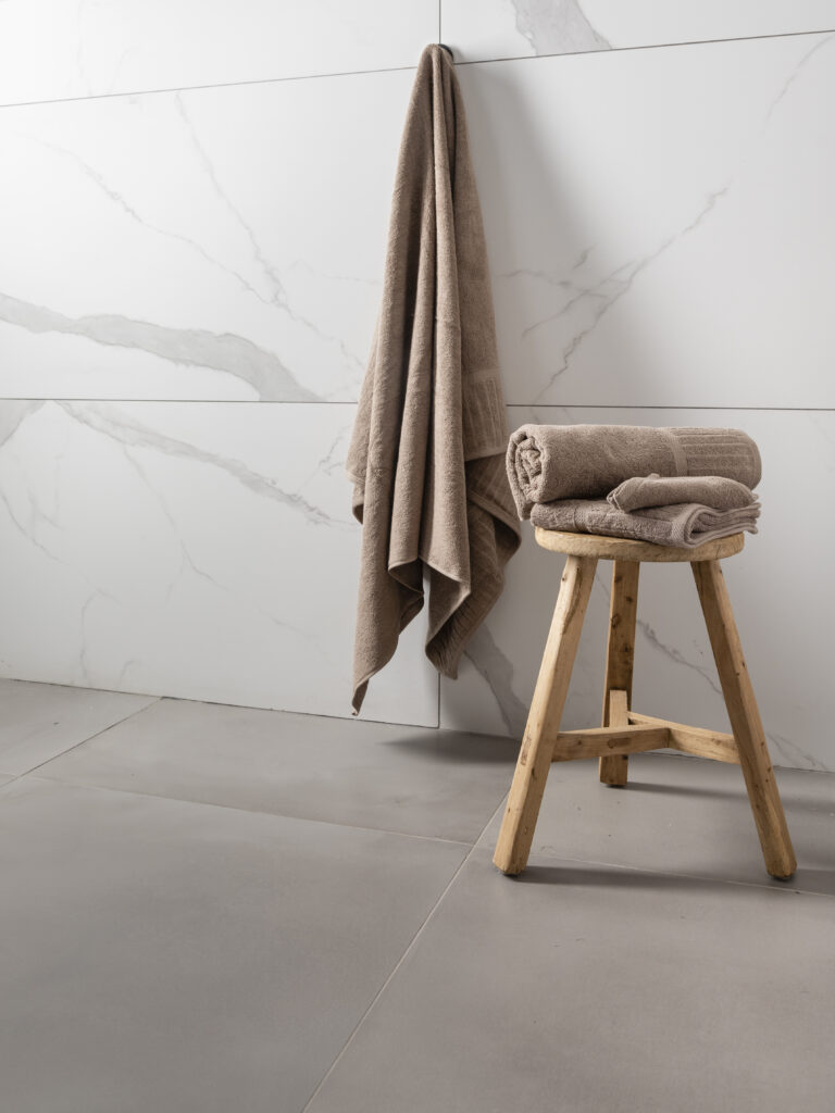 Luxury towels displayed in a modern bathroom, with a hanging bath towel and neatly folded matching towels arranged on a natural wooden stool against a white marble-effect tiled wall.