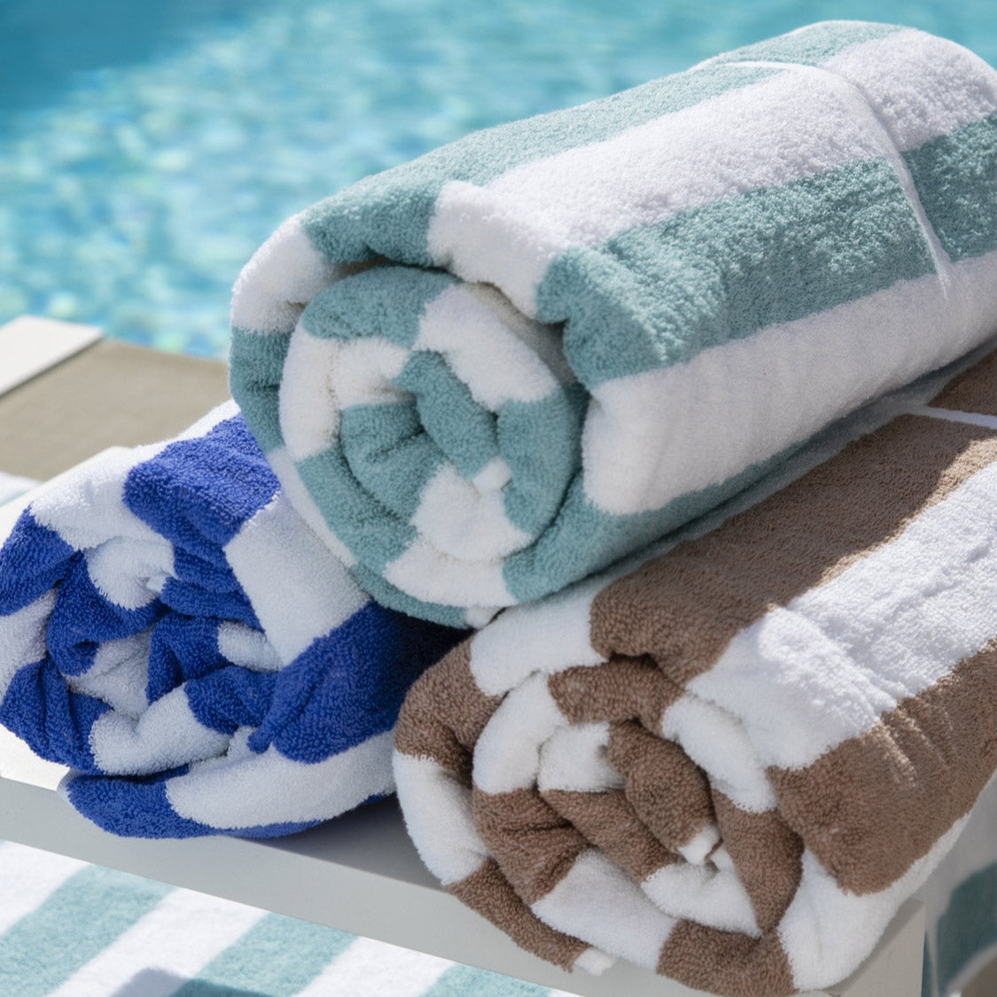 stripedbeachtowels-1 A collection of folded pool and beach towels in various colors, placed on a white rack by a poolside.